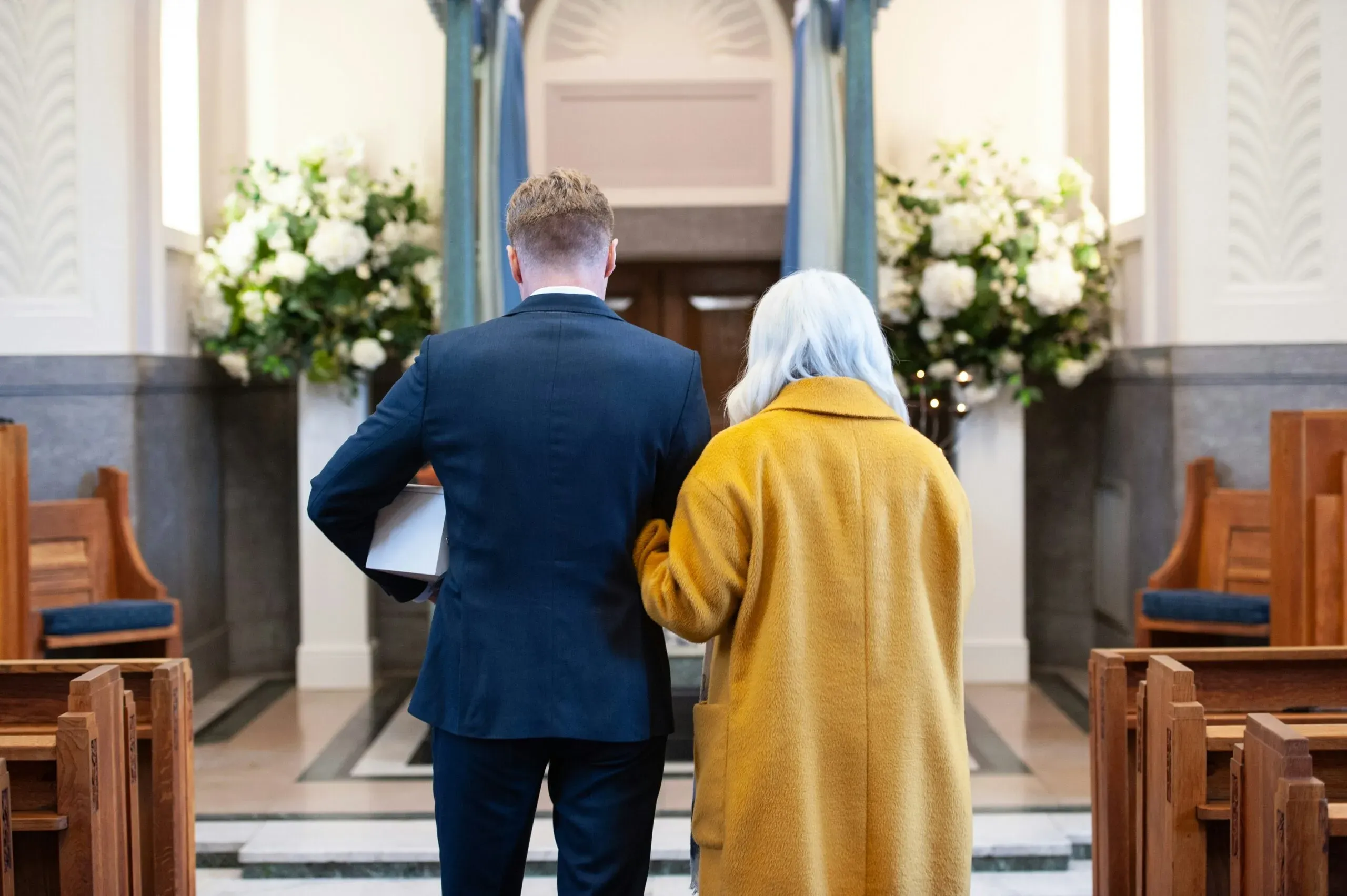 Couple endeuillé vu de dos, l'homme portant une urne funéraire dans une chapelle.